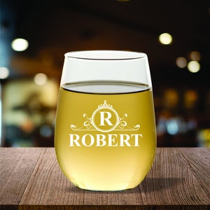 May include: A stemless wine glass filled with a light yellow liquid, likely wine. The glass has a white decorative design with the letter "R" inside a circle, and the name "ROBERT" below it. The glass sits on a wooden surface.