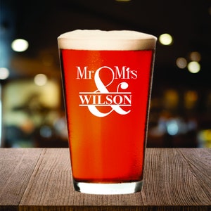 May include: A pint glass filled with a reddish-brown beverage and topped with white foam. The glass has the words "Mr & Mrs WILSON" in white script. The glass sits on a wooden surface.