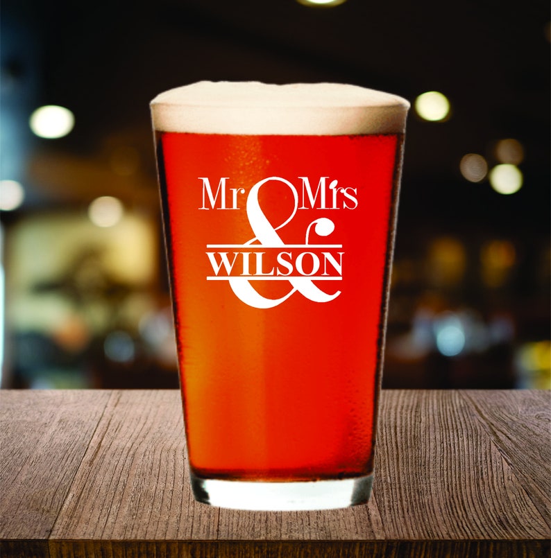 May include: A pint glass filled with a reddish-brown beverage and topped with white foam. The glass has the words "Mr & Mrs WILSON" in white script. The glass sits on a wooden surface.