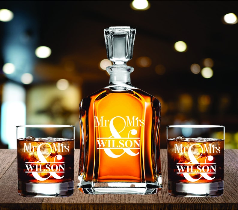May include: A personalized whiskey decanter and two matching glasses, all engraved with "Mr & Mrs WILSON". The decanter is filled with amber liquid and has a glass stopper. The glasses are filled with ice and a dark beverage.