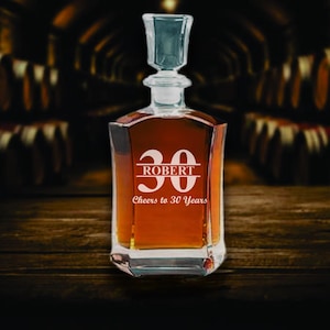 May include: A square-shaped glass decanter filled with amber liquid, featuring a crystal stopper. The front is etched with the number "30", the name "ROBERT", and the phrase "Cheers to 30 Years". The decanter sits on a dark wooden surface.
