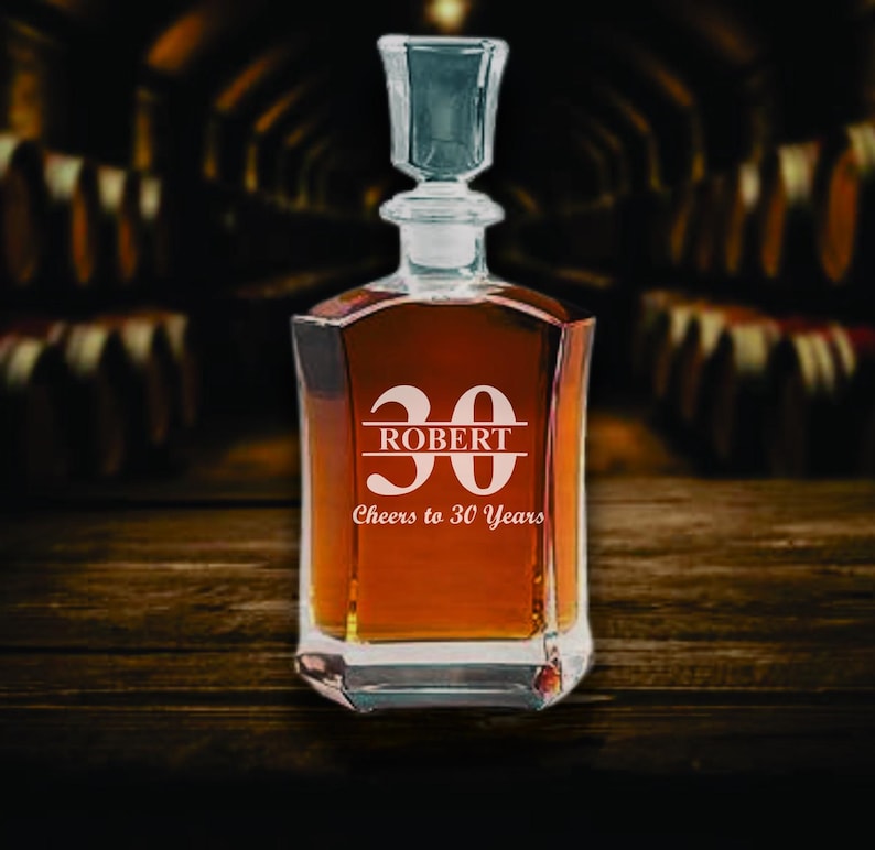 May include: A square-shaped glass decanter filled with amber liquid, featuring a crystal stopper. The front is etched with the number "30", the name "ROBERT", and the phrase "Cheers to 30 Years". The decanter sits on a dark wooden surface.