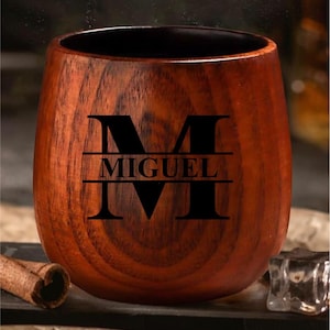 May include: A wooden cup with a dark interior and a natural wood grain exterior. The cup features the name "MIGUEL" in a black, bold font, with a large letter "M" above and below the name. The cup is designed for beverages.