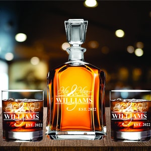 May include: A clear glass decanter and two matching lowball glasses filled with amber liquid and ice. The decanter and glasses are engraved with "Mr & Mrs WILLIAMS EST. 2022" in white script. The set is displayed on a wooden surface.