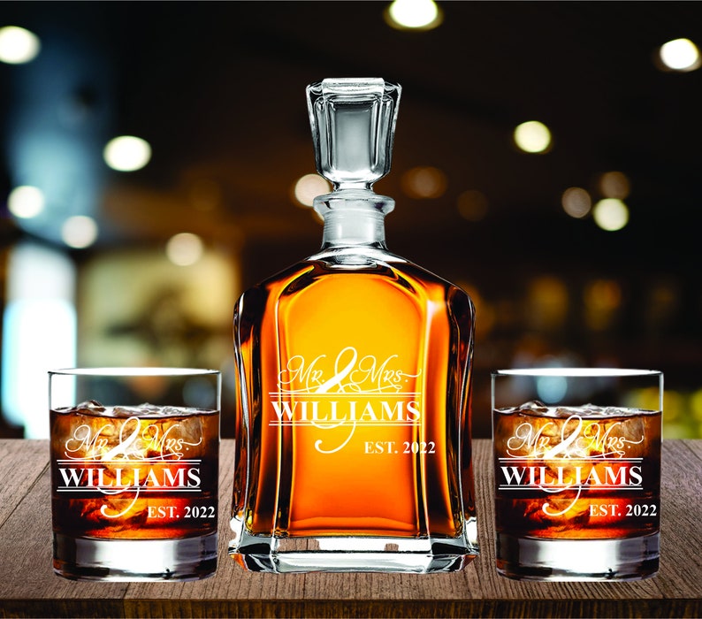 May include: A clear glass decanter and two matching lowball glasses filled with amber liquid and ice. The decanter and glasses are engraved with "Mr & Mrs WILLIAMS EST. 2022" in white script. The set is displayed on a wooden surface.