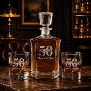 May include: A personalized whiskey decanter set with two matching glasses. The decanter and glasses are engraved with the name "ROBERT" and the number "50", along with the phrase "Cheers to 50 Years". The set is filled with amber-colored liquid.