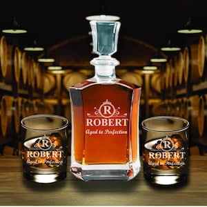 May include: A personalized decanter set with two matching glasses. The decanter and glasses are engraved with the name "ROBERT" and the phrase "Aged to Perfection". The set is displayed on a wooden surface.
