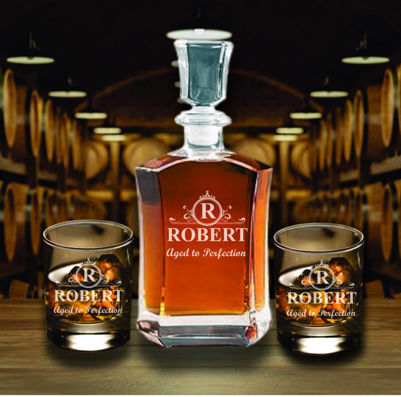 May include: A personalized decanter set with two matching glasses. The decanter and glasses are engraved with the name "ROBERT" and the phrase "Aged to Perfection". The set is displayed on a wooden surface.