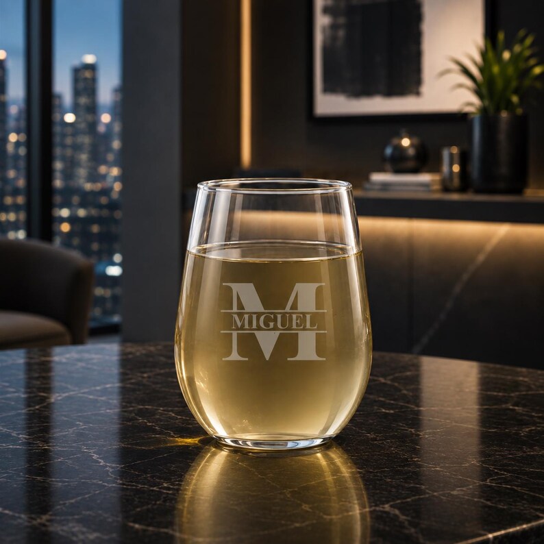 May include: A stemless wine glass filled with a light-colored liquid, possibly wine or juice. The glass is engraved with the name "MIGUEL" and a stylized initial "M". The glass sits on a dark, textured surface.