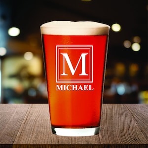 May include: A pint glass filled with a reddish-brown beverage and topped with white foam. The glass features a white square with the letter "M" and the name "MICHAEL" in a serif font.