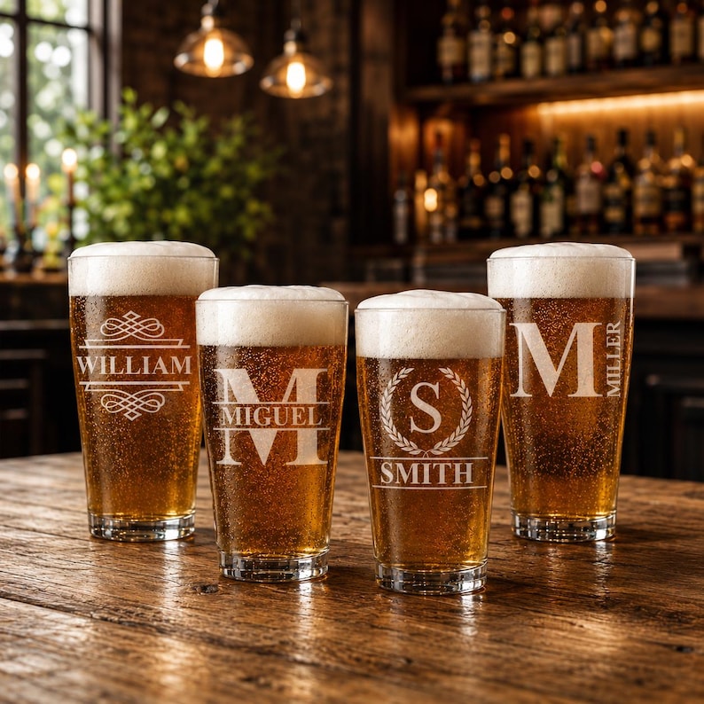 May include: Four clear pint glasses filled with beer and topped with white foam, each etched with a different name: William, Miguel, Smith, and Miller. The glasses sit on a wooden table in a bar setting, illuminated by overhead lights.