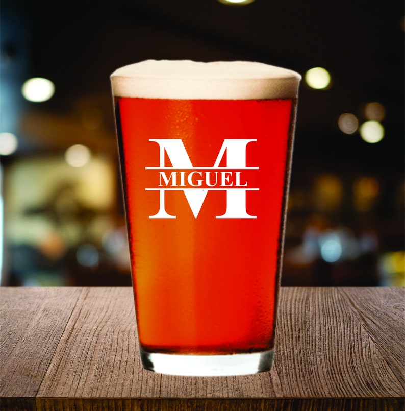 May include: A pint glass filled with a reddish-brown beverage and topped with white foam. The glass has a white monogram with the letter "M" and the name "MIGUEL" printed in a sans-serif font.