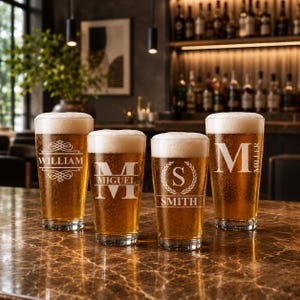 May include: Four pint glasses filled with beer and topped with white foam. Each glass is engraved with a different name or initial: "WILLIAM", "MIGUEL", "S SMITH", and "M MILLER". The glasses are on a brown marble surface.