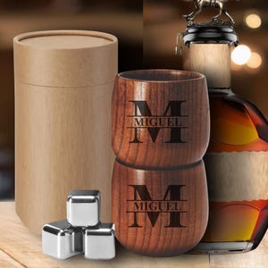 May include: A set of two wooden tumblers with the name "MIGUEL" engraved, stacked on top of each other. The tumblers are made of dark wood and have a natural finish. Also included are stainless steel ice cubes and a cylindrical cardboard container.