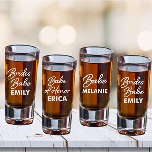 May include: Four clear glass shot glasses with the text "Brides Babe Emily", "Babe of Honor Erica", "Babe Melanie", and "Brides Babe Emily" etched on them.