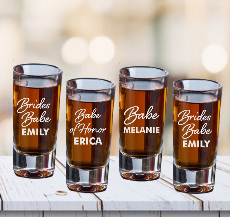 May include: Four clear glass shot glasses with the text "Brides Babe Emily", "Babe of Honor Erica", "Babe Melanie", and "Brides Babe Emily" etched on them.