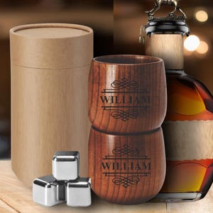 May include: A set of two wooden whiskey glasses with the name "WILLIAM" engraved on them, stacked on top of each other. The glasses are next to a brown cardboard tube, stainless steel ice cubes, and a bottle of liquor.