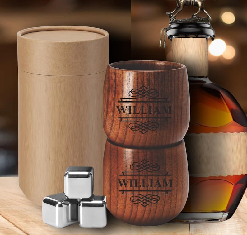 May include: A set of two wooden whiskey glasses with the name "WILLIAM" engraved on them, stacked on top of each other. The glasses are next to a brown cardboard tube, stainless steel ice cubes, and a bottle of liquor.