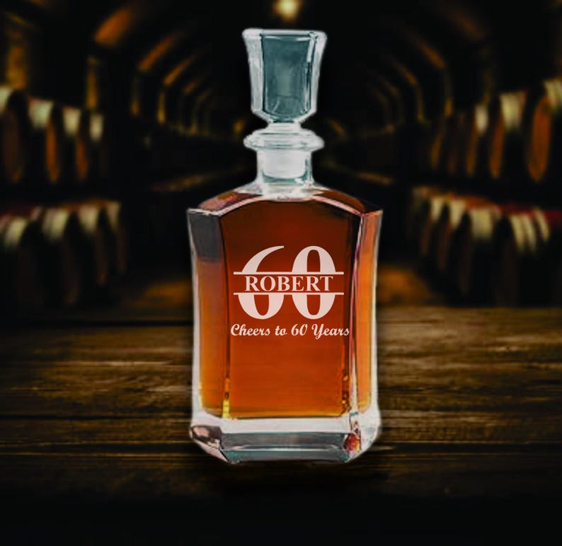 May include: A clear glass decanter filled with amber liquid, featuring a glass stopper. The decanter is engraved with the number "60", the name "ROBERT", and the phrase "Cheers to 60 Years". The decanter sits on a wooden surface.