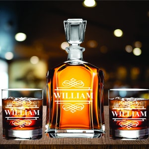 May include: A personalized whiskey decanter set with two matching glasses. The decanter and glasses are engraved with the name "WILLIAM" in a decorative font. The set is filled with amber-colored liquid and ice cubes, displayed on a wooden surface.