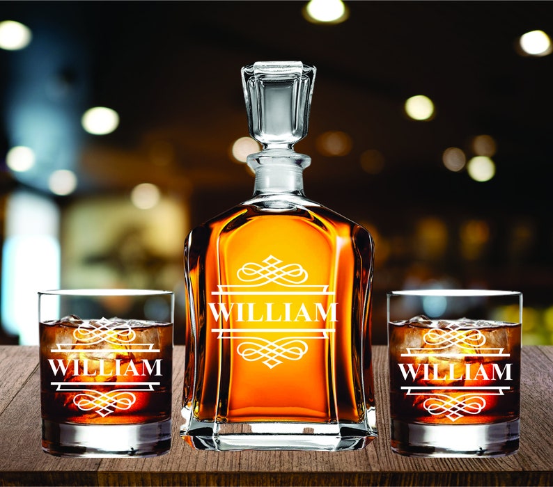 May include: A personalized whiskey decanter set with two matching glasses. The decanter and glasses are engraved with the name "WILLIAM" in a decorative font. The set is filled with amber-colored liquid and ice cubes, displayed on a wooden surface.