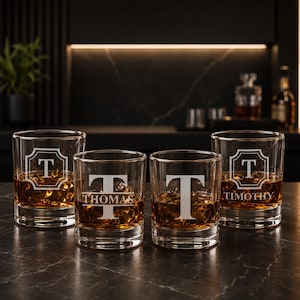 May include: Four clear whiskey glasses with ice and amber liquid. Each glass is engraved with a capital "T" and a name. Two glasses have the name "Thomas" and "Timothy". The glasses are on a dark surface.