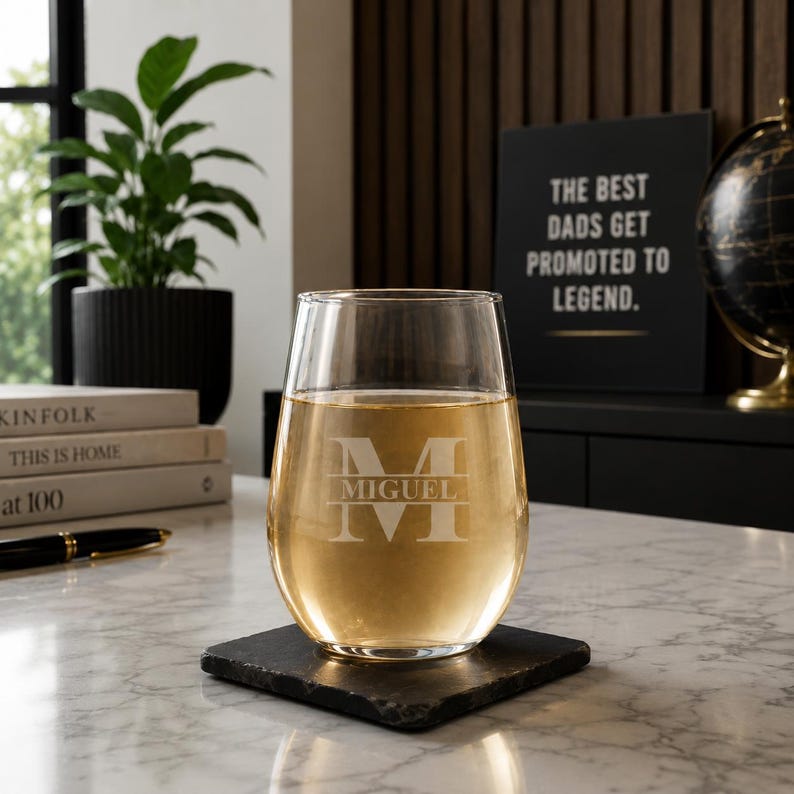 May include: A stemless wine glass filled with a light-colored liquid, resting on a square black coaster. The glass is etched with the name "MIGUEL" and a large initial "M". A sign in the background reads "THE BEST DADS GET PROMOTED TO LEGEND."
