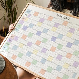 Könnte beinhalten: Ein großer, gerahmter 2026-Plan-Kalender mit Rasterlayout. Der Kalender zeigt pastellfarbene Blöcke für jeden Monat und Tag. Der Rahmen besteht aus hellem Holz. Der Kalender wird von einer Person gehalten.