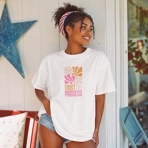 Op de afbeelding: Een wit t-shirt met korte mouwen voorzien van een grafisch ontwerp met vier gestileerde bloemvormen in roze, oranje en crèmetinten, geplaatst boven de tekst "TRUST THE PROCESS" gedrukt in een vet, schreefloos lettertype in oranje en roze.