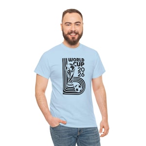 Puede incluir: Una camiseta de manga corta de color azul claro con un estampado gráfico negro en el pecho. El diseño incluye el texto "WORLD CUP 2026" junto a una ilustración estilizada de un trofeo y un balón de fútbol, enmarcado por líneas verticales.