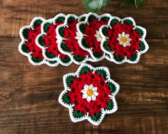 Peut inclure: Ensemble de six dessous-de-verre en crochet rouge, blanc et vert en forme de poinsettia. Les dessous-de-verre sont en forme de fleur avec un centre blanc et des feuilles vertes.