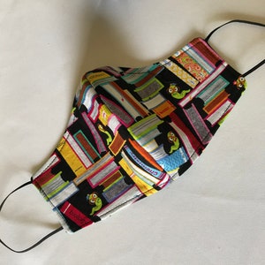 Op de afbeelding: Een zwart gezichtsmasker met een kleurrijke print van gestapelde boeken en kleine groene uilen. De boeken hebben titels zoals "biologie", "chemie" en "geschiedenis".