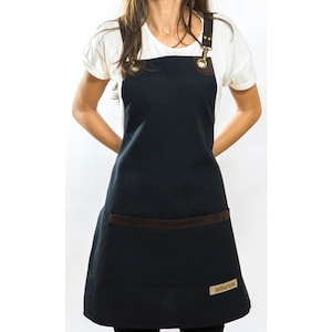 May include: Black apron with brown leather trim and adjustable straps. The apron has a large pocket on the front and a small logo on the bottom right corner.