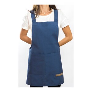 Peut inclure: Un tablier en denim bleu avec des bretelles réglables et une grande poche sur le devant. Le tablier a un patch en cuir brun avec le mot "Studio" dessus.