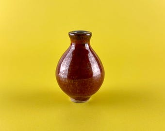 May include: A small, round, brown ceramic vase with a dark rim and a light-colored base. The vase has a glossy finish and sits against a bright yellow background. The vase is likely used for decorative purposes.