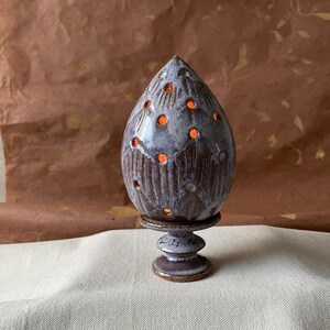 Pode incluir: Um queimador de incenso de cerâmica em forma de lágrima com um esmalte branco e detalhes marrons. O queimador tem uma base com um design de pedestal.