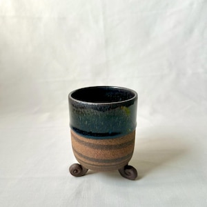 May include: A handmade ceramic cup with a dark blue and green glazed top and a brown, textured base. The cup rests on three small, curled feet. This unique piece is ideal for holding small plants or as a decorative item.