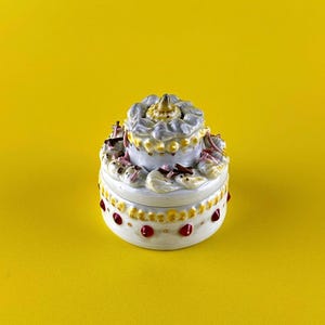 May include: A two-tiered, white ceramic trinket box with gold accents and red jewels. The top tier has a decorative, cloud-like design. The box is set against a bright yellow background, creating a striking contrast.