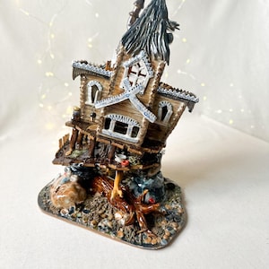 May include: A whimsical ceramic house with a brown wooden exterior, a blue roof, and white trim. The house is perched on a tree trunk and surrounded by rocks and pebbles.