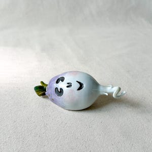May include: A whimsical ceramic figurine of a smiling radish. The radish has a white body with a purple top and green leaves. The face features black eyes and a curved mouth. The radish has a curly white tail.