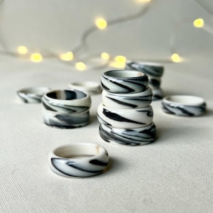 May include: A collection of white and black swirl-patterned rings. The rings are stacked and scattered, showcasing a marbled design. The rings are made of a glossy material and are set against a soft, neutral background.