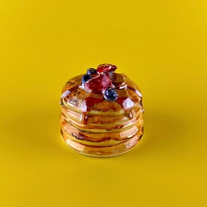May include: A ceramic stack of pancakes with syrup, blueberries, and a strawberry on top. The pancakes are a golden brown color, and the syrup is a rich, dark brown. The background is a solid yellow.