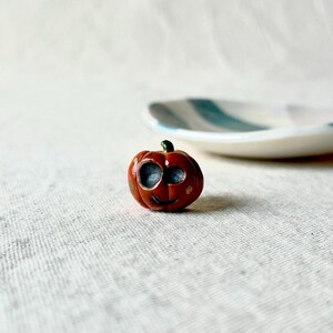May include: A small orange ceramic pumpkin with a black painted face and gold dots. The pumpkin is sitting on a white fabric surface.