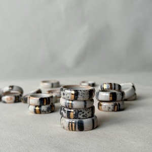 May include: Ceramic rings with a black and white speckled pattern and gold stripes. Some rings are stacked, others scattered. The rings are on a neutral surface. The design is minimalist and modern.