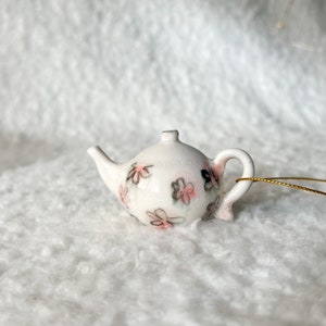 May include: A small white teapot with a floral design in pink and gray. The teapot has a gold string attached to the handle.