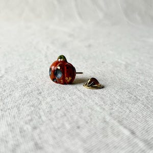 May include: A small, orange pumpkin-shaped pin with a green stem and blue eyes. The pin has a gold-colored backing and is set against a neutral background.