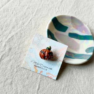 May include: A small, orange ceramic pumpkin with white speckles and a green stem. The pumpkin is on a white card with the text "Mist Ceramics Handmade in LA, CA".