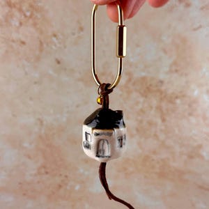 Puede incluir: Un llavero de mosquetón dorado con un pequeño colgante de casa de cerámica. La casa tiene un tejado negro, paredes blancas y pequeñas ventanas. Un cordón de cuero marrón y una cuenta dorada conectan la casa al llavero.