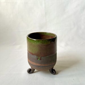 Puede incluir: Una taza pequeña de cerámica marrón con un esmalte verde y tres patas. La taza está sobre una superficie blanca.