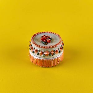 May include: A small, round ceramic trinket box with a lid. The box is decorated with red roses on top and floral patterns around the sides. The base is orange, and the lid is light gray. The background is a solid yellow.
